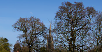 The landscape photograph shows St James' Church in Louth, Lincolnshire, with its prominent spire rising behind leafless trees. The image was taken in the early afternoon during spring, as indicated by the presence of budding foliage and clear blue sky. The moon is visible in the sky above the church, creating a contrast between natural and architectural features. St James' Church is a notable landmark in Louth, Lincolnshire, and its spire is a distinctive element within the town. The composition highlights both the church and seasonal changes typical of spring in this region.